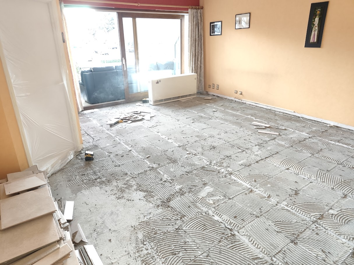 Sol de salon en cours de rénovation, ancien carrelage retiré et support préparé avant pose d’un nouveau revêtement, avec baie vitrée en arrière-plan.