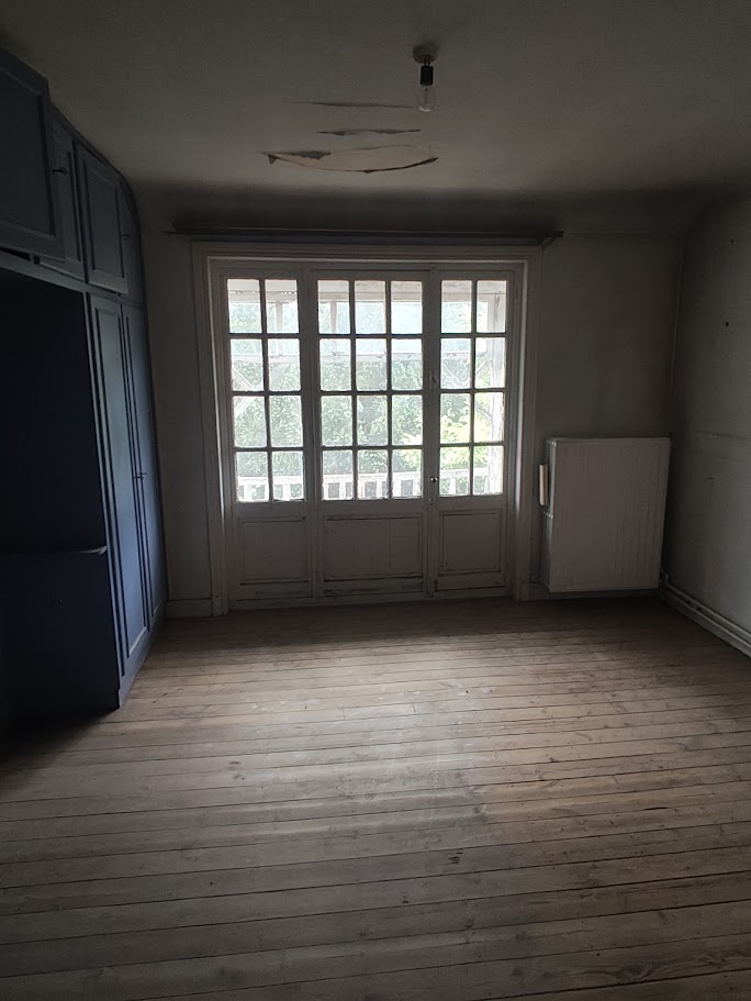 An empty room with a large window and wooden floor, featuring a radiator and cabinet on the right wall, and a closet on th...