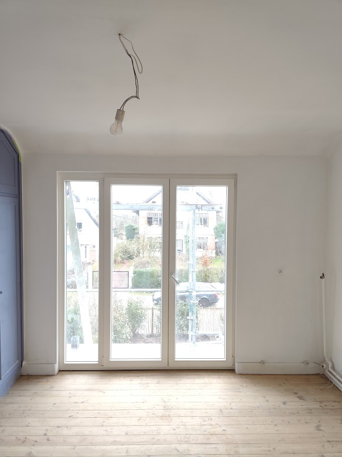 A room with white walls, a lightbulb hanging from the ceiling, and a large window with a view of the outside.