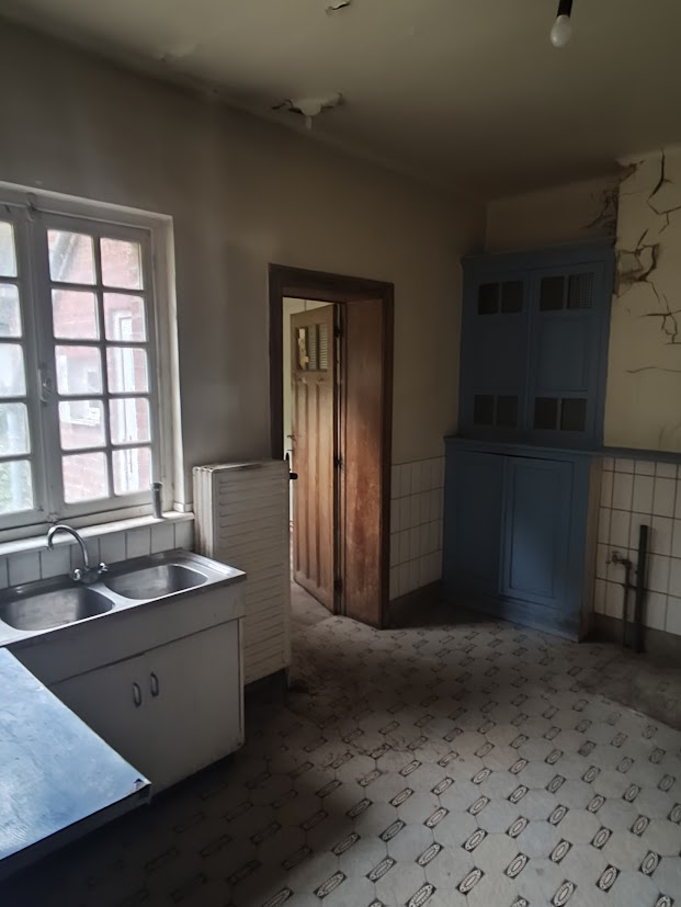 An abandoned kitchen with white walls, a double sink, and a large window.